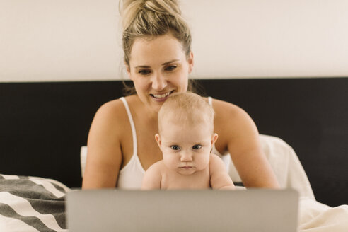 Baby-Mädchen und Mutter sitzen im Bett und starren auf den Laptop - CUF02800