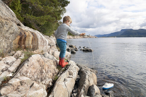 Junge steht auf einem Felsen am Fjord und spielt mit einem Spielzeugboot, Aure, More og Romsdal, Norwegen - CUF03013