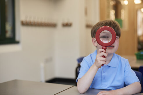 Schuljunge, der durch ein Vergrößerungsglas in einem Klassenzimmer in der Grundschule schaut, Porträt - CUF03073