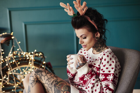 Junge Frau sitzt zu Hause, trägt Weihnachtspulli und Geweih, trinkt ein heißes Getränk, lizenzfreies Stockfoto