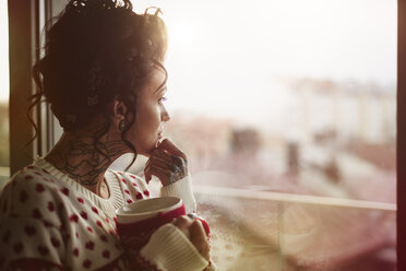 Junge Frau schaut aus dem Fenster, hält ein heißes Getränk in der Hand, nachdenklicher Ausdruck - CUF03123