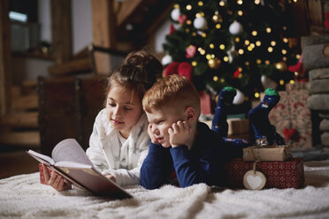 Junges Mädchen und Junge entspannen sich neben dem Weihnachtsbaum und lesen ein Buch - CUF03132