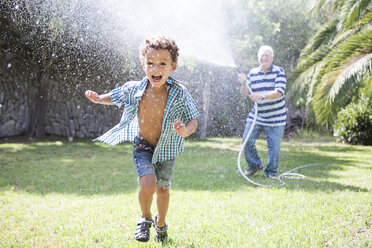 Boy running away from grandfather spraying hosepipe in garden - CUF03174