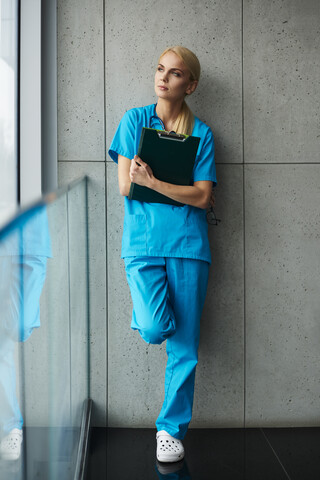 Porträt eines Chirurgen, an die Wand gelehnt, Klemmbrett in der Hand, nachdenklicher Ausdruck, lizenzfreies Stockfoto