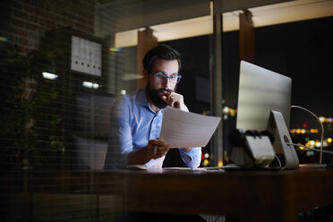 Young businessman reading paperwork at office desk at night - CUF03298