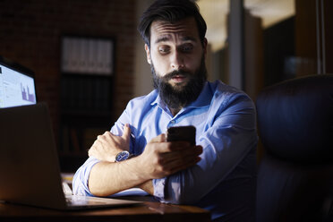 Junger Geschäftsmann schaut nachts auf sein Smartphone am Schreibtisch im Büro - CUF03308