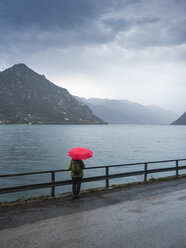 Italien, Lombardei, Rückenansicht einer älteren Frau mit Rucksack und rotem Regenschirm mit Blick auf den Idrosee - LAF02028