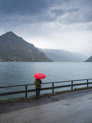 Italy, Lombardy, back view of senior woman with backpack and red umbrella looking at Lake Idro - LAF02028