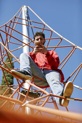 Young man using cell phone on a climbing frame on a playground - JRFF01640