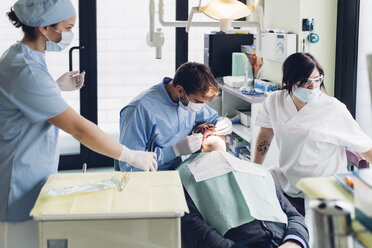 Dentist looking into male patient's mouth, dental nurses preparing equipment - CUF03445