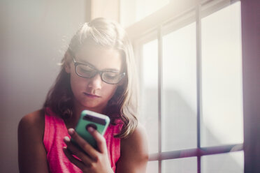 Young girl wearing glasses, beside window, using smartphone - CUF03512