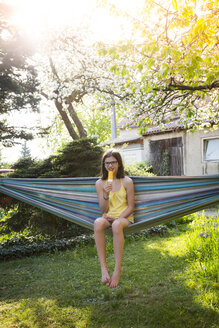 Lächelndes Mädchen sitzt auf einer Hängematte im Garten und isst einen Eislutscher - LVF06960
