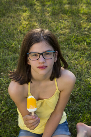 Porträt eines Mädchens mit Brille, das auf einer Wiese sitzt und einen Eislutscher isst, lizenzfreies Stockfoto