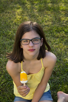 Porträt eines Mädchens mit Brille, das auf einer Wiese sitzt und einen Eislutscher isst - LVF06961