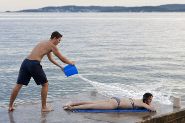 Junger Mann wirft Eimer mit Wasser über eine Sonnenbadende, Orebic, Kroatien - CUF03822