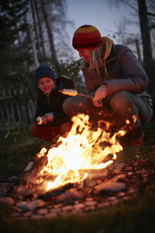 Ältere Frau und Sohn rösten Marshmallows am Lagerfeuer im Garten in der Abenddämmerung - CUF03838