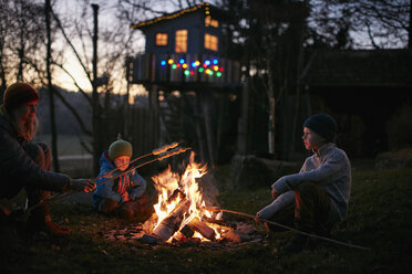 Mature woman and two sons toasting marshmallows on campfire at night - CUF03845