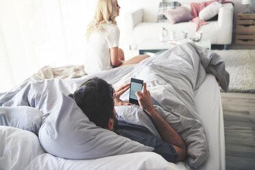 Man in bed using smartphone, woman sitting at end of bed - CUF03945