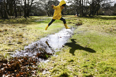 Boy in yellow anorak jumping over puddle in park - CUF03999