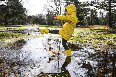 Boy in yellow anorak splashing in park puddle - CUF04033