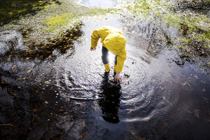 Junge im gelben Anorak beugt sich in einer Parkpfütze nach vorne - CUF04034