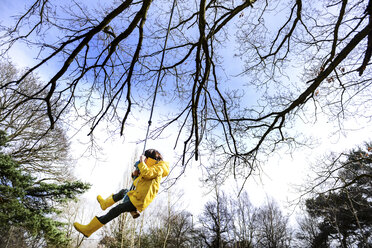 Boy in yellow anorak swinging from park tree - CUF04035