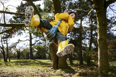 Boy in yellow anorak swinging from park tree - CUF04036