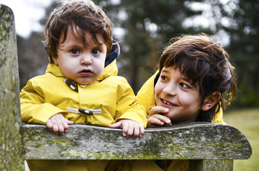 Portrait of baby boy and big brother in yellow anoraks on park bench - CUF04039
