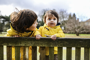 Portrait of baby boy and big brother in yellow anoraks on park bench - CUF04040