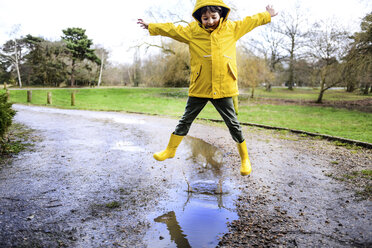 Boy in yellow anorak jumping above puddle in park - CUF04043