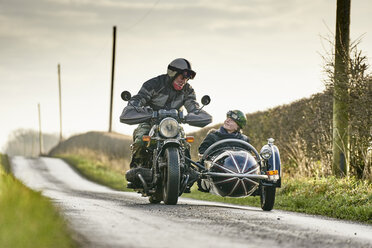 Ein älterer Mann und sein Enkel fahren mit Motorrad und Beiwagen eine Landstraße entlang - CUF04150