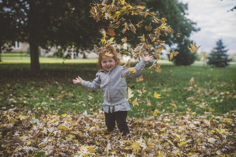 Porträt eines rothaarigen weiblichen Kleinkindes im Park, das Herbstblätter wirft, lizenzfreies Stockfoto