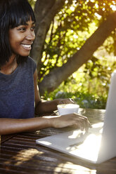 Junge Frau sitzt im Freien, benutzt einen Laptop und trinkt Kaffee - CUF04450