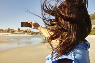Mature woman on beach, taking selfie, using smartphone, rear view - CUF04454