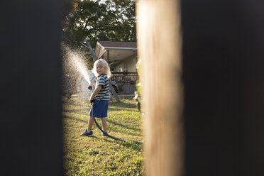 Blick durch eine Lücke im Zaun auf einen Jungen, der Wasser aus einem Schlauch versprüht - ISF00945
