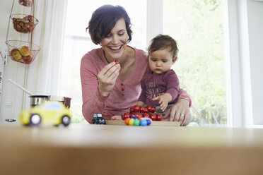 Mother holding baby girl at kitchen counter, pile of tomatoes in front of them - ISF01028