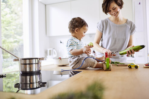 Baby girl sitting on kitchen counter watching mother slicing cucumber - ISF01031