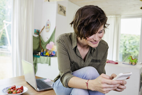 Mid adult woman sitting on kitchen table, using smartphone - ISF01037