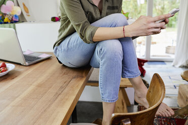 Mid adult woman sitting on kitchen table, using smartphone, low section - ISF01038