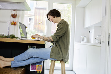 Mittlere erwachsene Frau in der Küche sitzend, mit Laptop, lachend - ISF01039