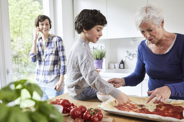 Großmutter und Enkel beim Pizzabacken in der Küche, Mutter im Hintergrund mit Smartphone - ISF01054