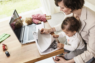 Mutter sitzt mit ihrer kleinen Tochter auf dem Tisch und sieht Papierkram durch, vor ihr steht ein Laptop - ISF01095