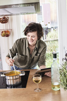 Mid adult woman stirring pot on stove in kitchen, holding digital tablet, laughing - ISF01099