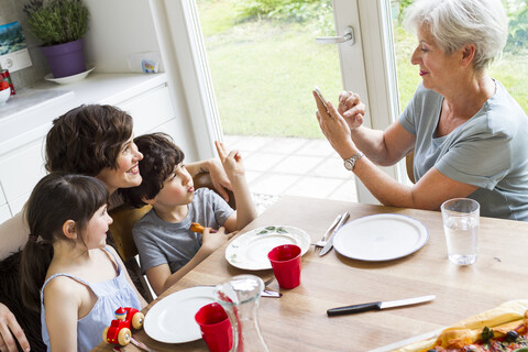 Großmutter sitzt in der Küche und fotografiert ihre erwachsene Tochter und Enkelkinder mit dem Smartphone, lizenzfreies Stockfoto