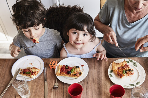 Großmutter sitzt mit Enkelkindern am Küchentisch und isst Pizza, Blick von oben, lizenzfreies Stockfoto