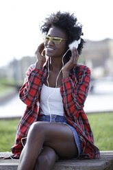 Porträt einer lächelnden jungen Frau, die auf einer Bank sitzt und mit Kopfhörern Musik hört - JSRF00043
