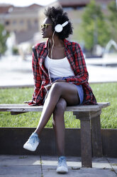 Porträt einer jungen Frau, die auf einer Bank im Stadtpark sitzt und mit Kopfhörern Musik hört - JSRF00047