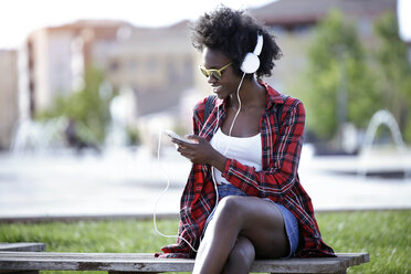 Lächelnde junge Frau sitzt auf einer Bank im Stadtpark und hört mit Kopfhörern Musik - JSRF00048