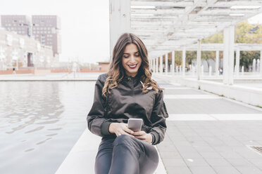 Young woman sitting outdoors, using smartphone, smiling - CUF04500