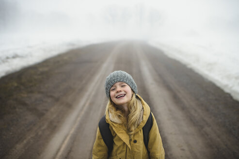 Portrait of girl in knit hat standing in middle of foggy dirt road laughing - CUF04539